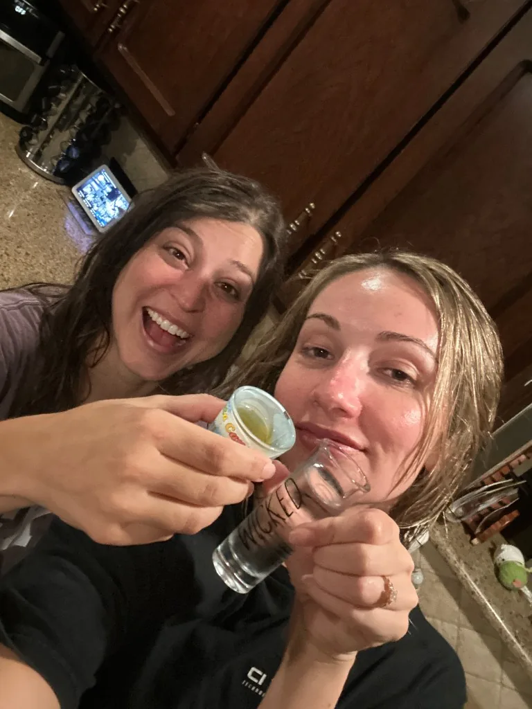 Two CI Web Group teammates laughing in a kitchen as they raise small glasses in a toast, one glass hand-labeled WICKED